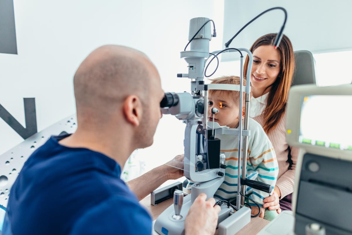 Optometrist performing an exam on a child with parent in background. 